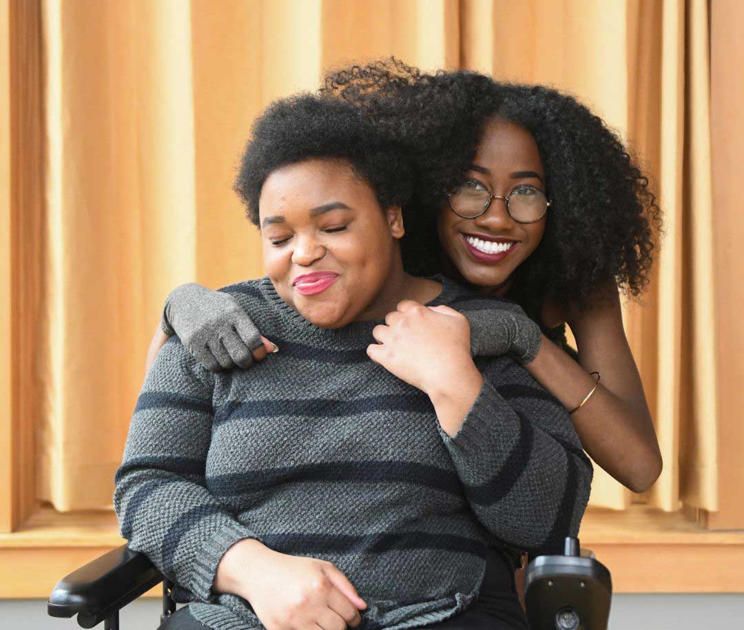 Woman in a wheelchair smiling with her eyes closed with a woman behind hugging her looking at the camera