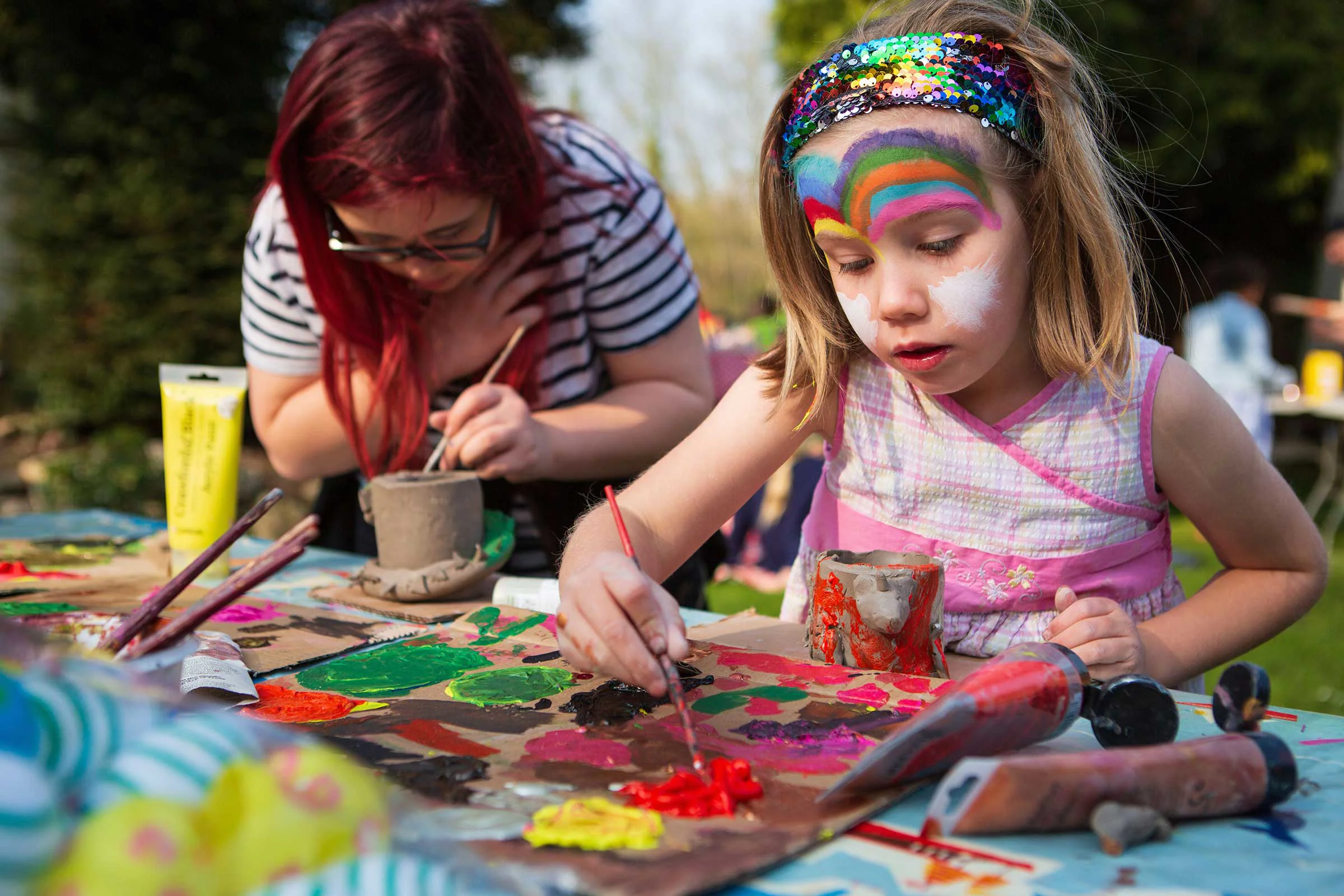 Child with face paint, painting a picture with an adult in the background making a pot