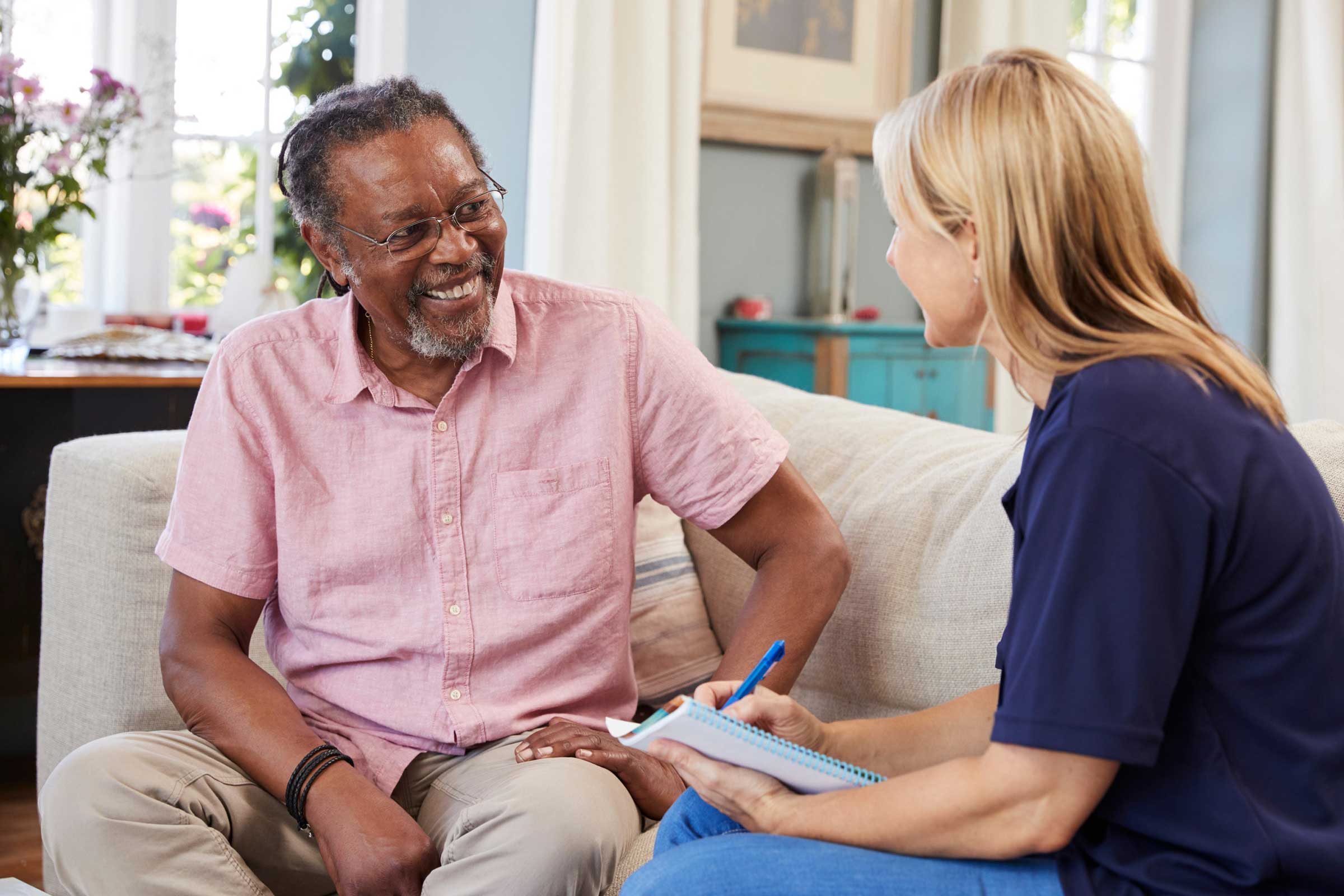 Photo focused on an older man, smiling, talking with a support worker at home