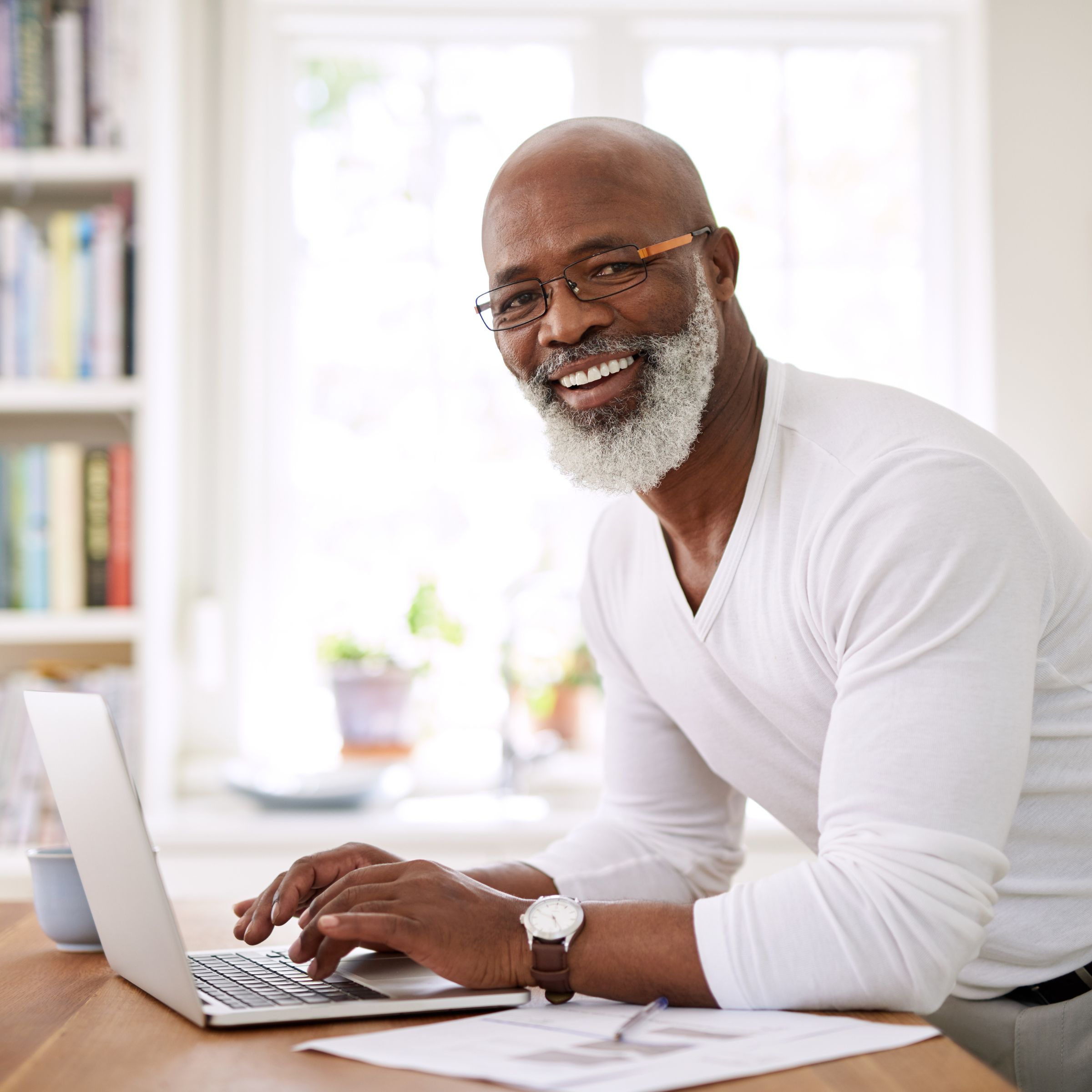 Man wearing a white top taps at his laptop whilst smiling towards the camera