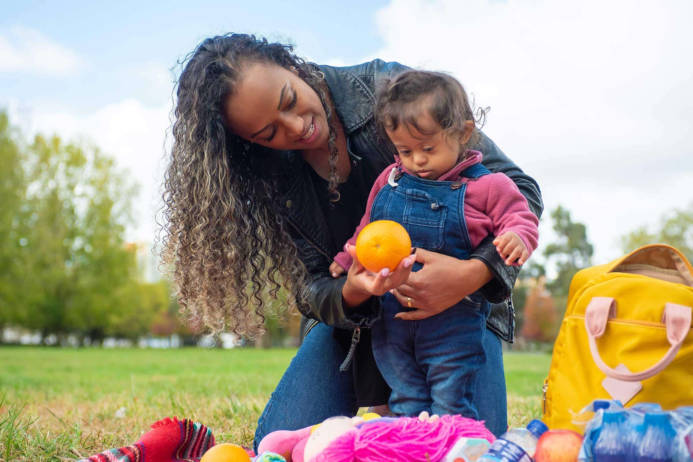 black women kneeling with her arm around a toddler and holding a orange for the toddler to take