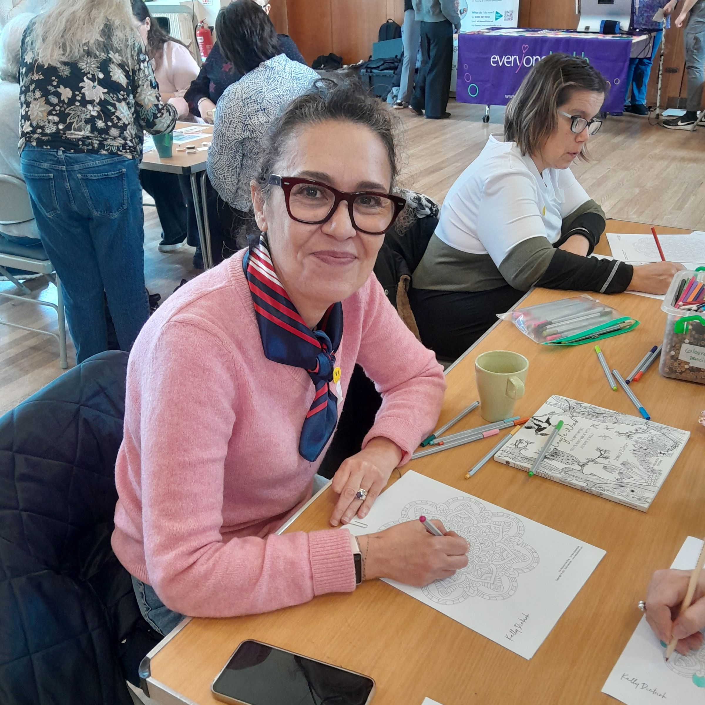 Woman wearing a pink jumper smiling at the camera whilst doing some colouring