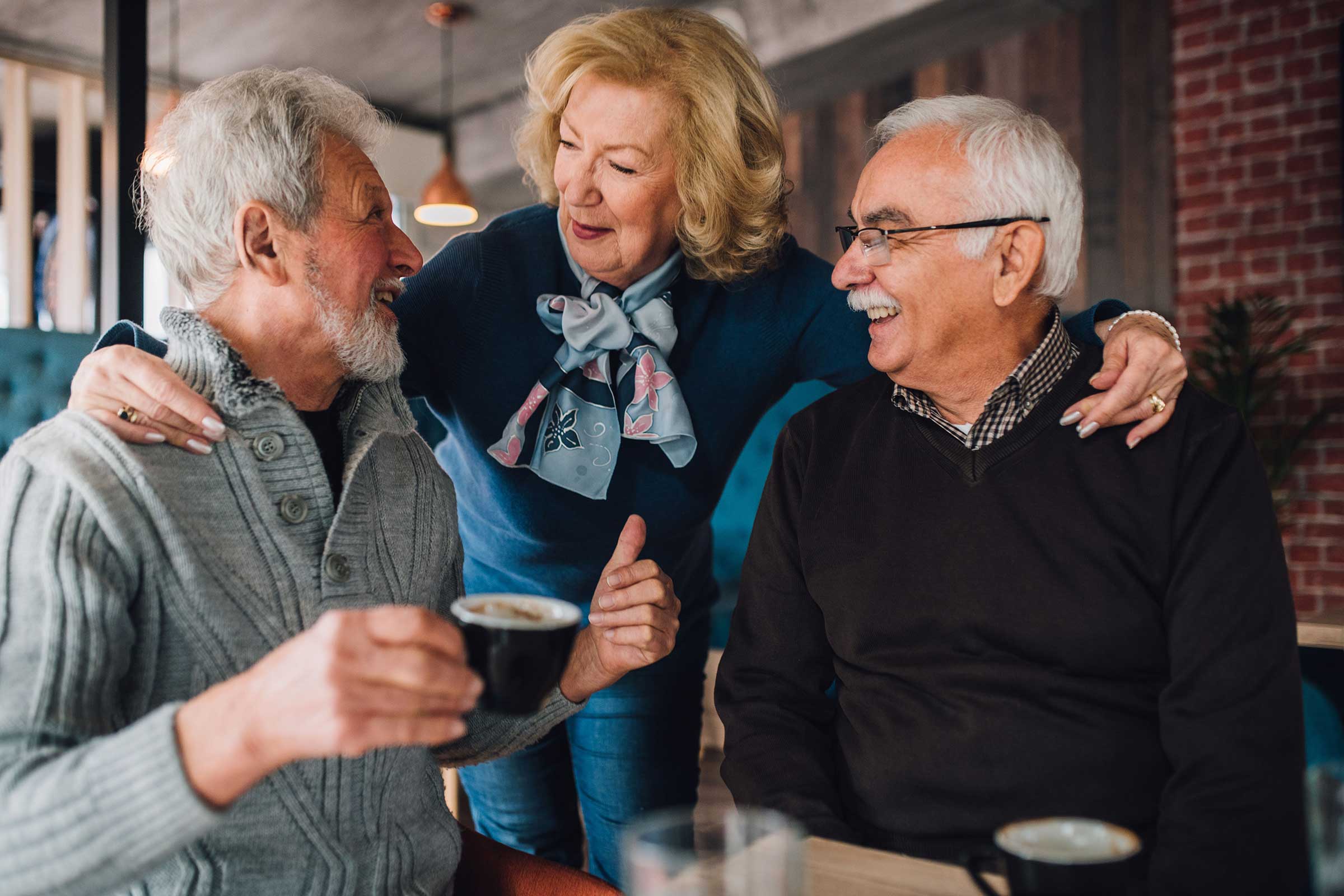 Two men sat down with a women stood between them with her arms around their shoulders talking to them